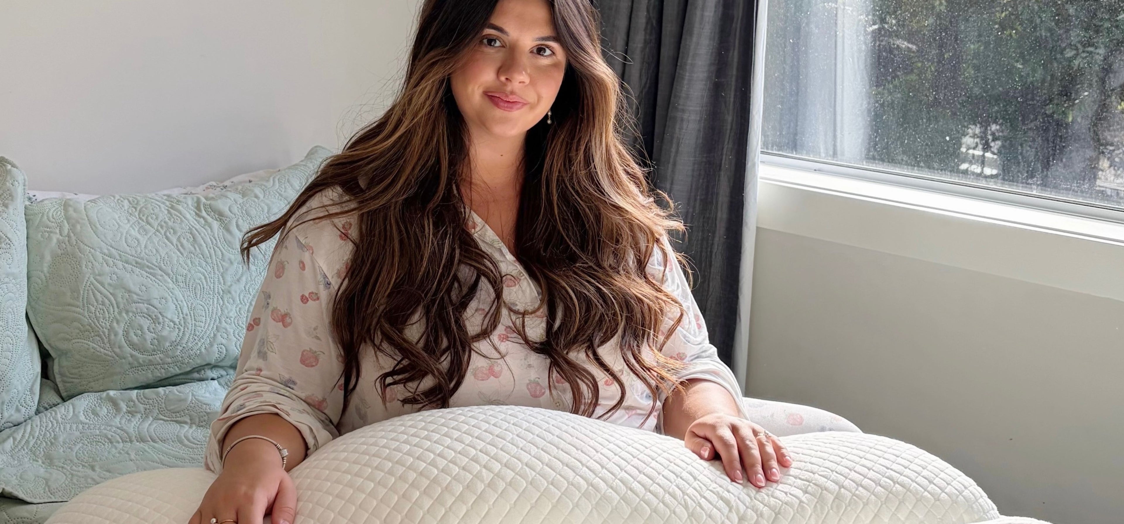 Woman sitting on a bed holding a pregnancy pillow in a bright bedroom