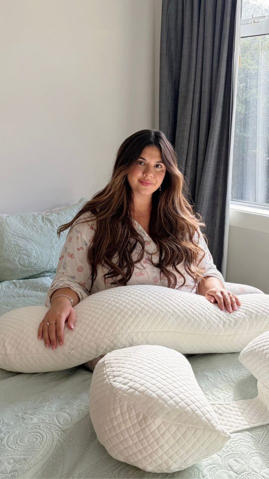 Woman sitting on a bed with a pregnancy pillow, surrounded by soft bedding.