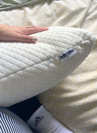 White quilted pillow with 'Belly Bliss' label held by a person, lying on a textured surface.