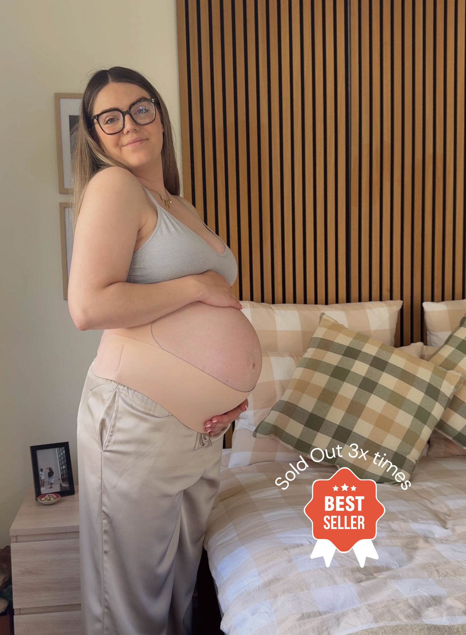 A pregnant woman wearing a beige maternity belly band and light gray tank top stands in a bedroom next to a bed with plaid pillows. A red 'Sold Out 3x times BEST SELLER' badge is overlaid in the bottom right corner.