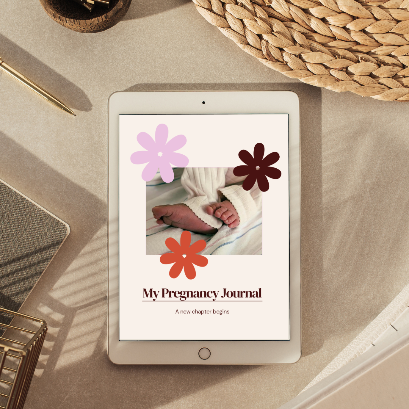 A tablet lying on a beige surface, displaying the cover of a pregnancy journal with the title 'My Pregnancy Journal' and decorative flower graphics. The scene is styled with a notebook, pen, woven mat, and basket in soft natural light.