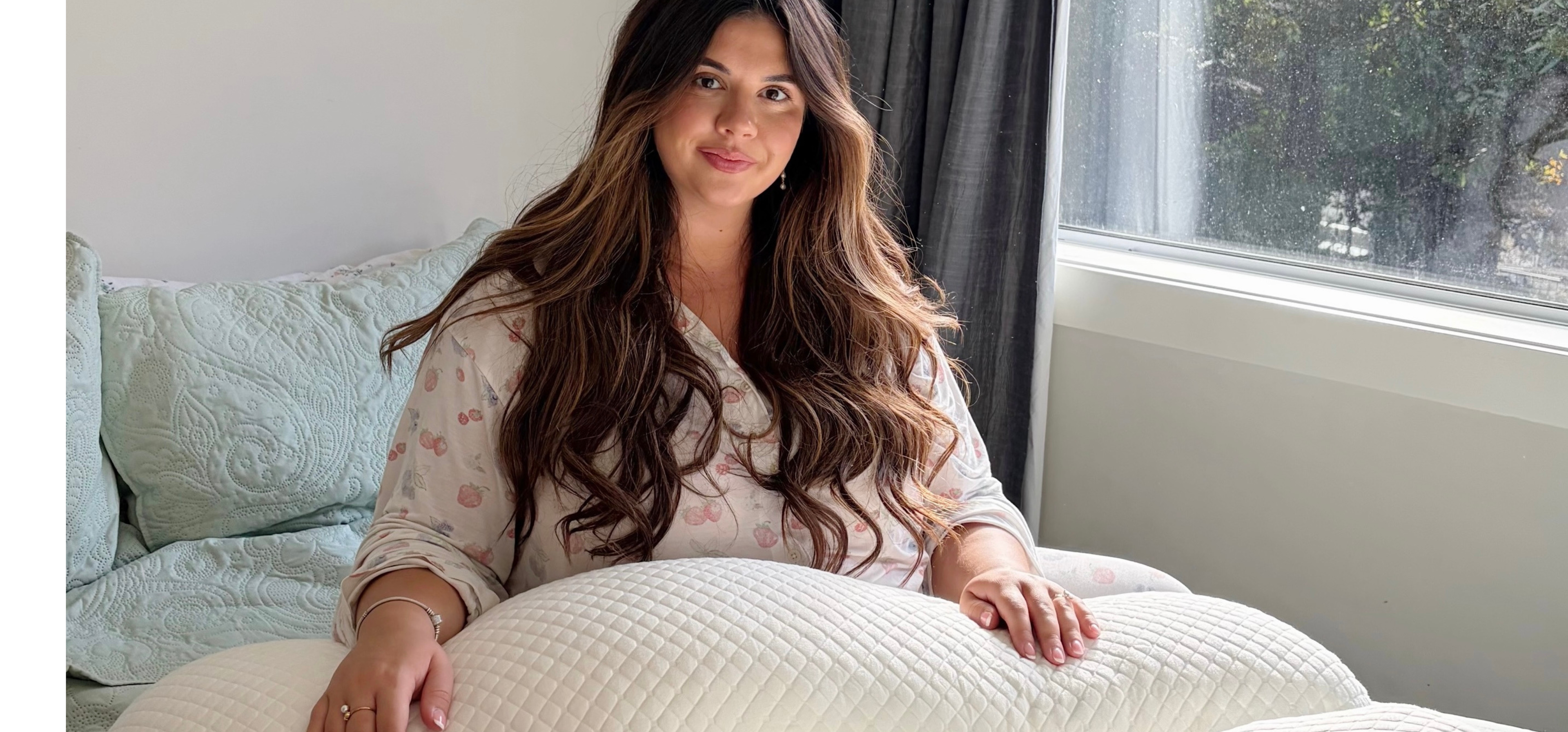 Woman sitting on a bed with a large white pregnancy pillow, smiling at the camera.