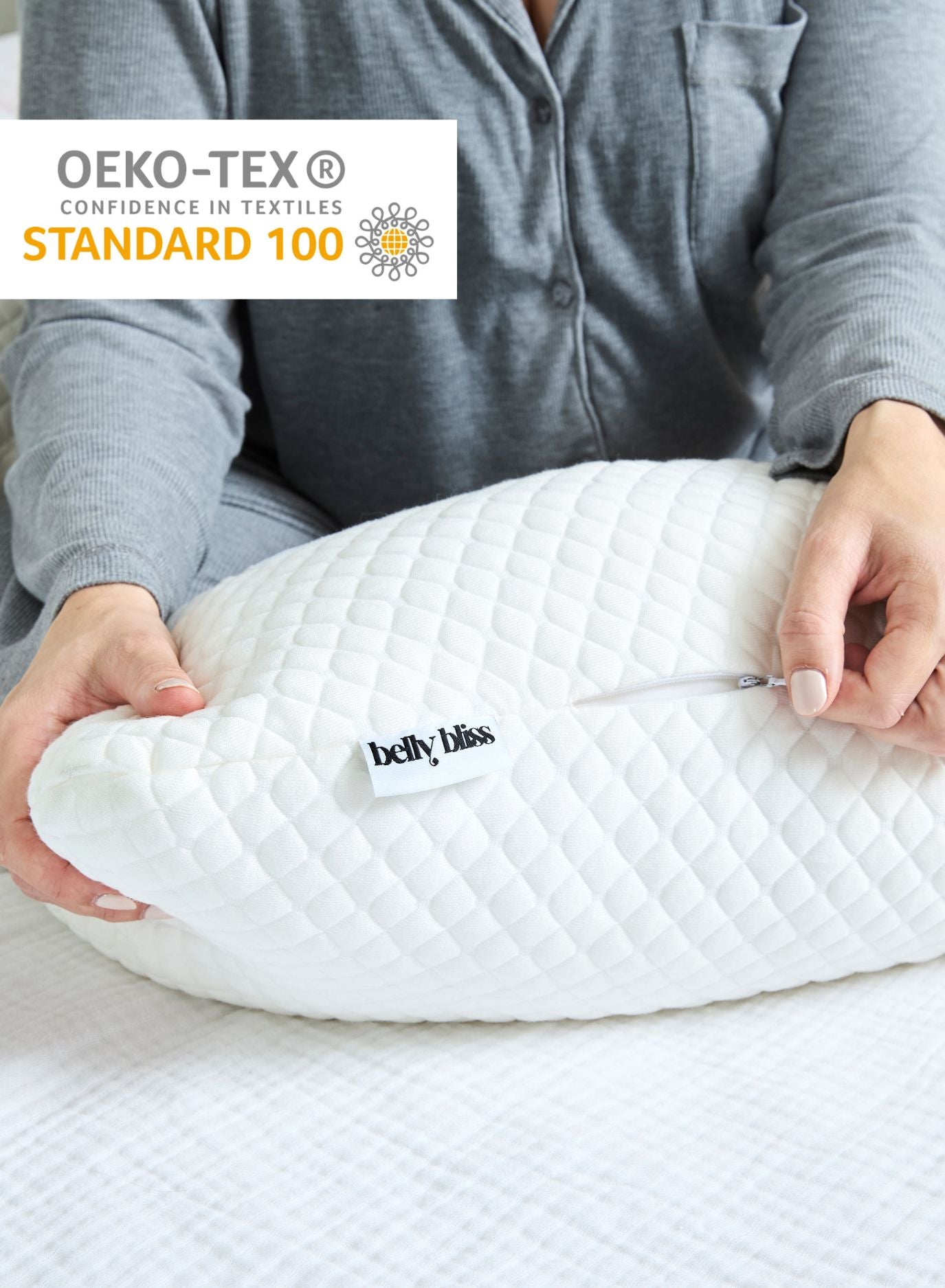 Person holding a white belly pillow with 'belly bliss' branding, OEKO-TEX Standard 100 logo visible.