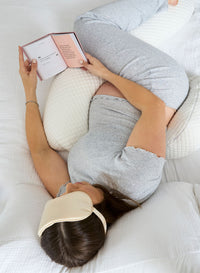 Person lying on a U-shaped body pillow reading a book with an eye mask on.