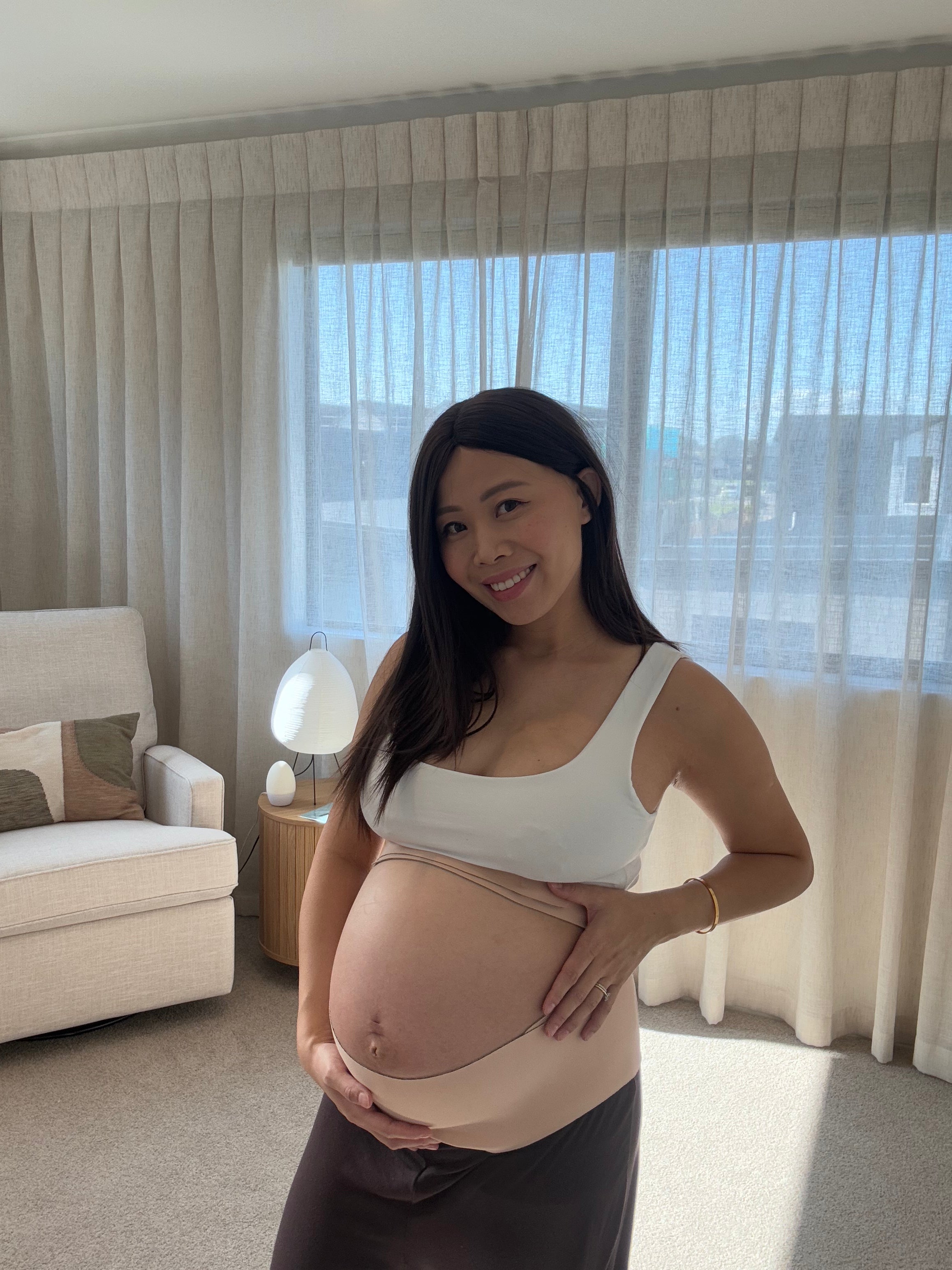 A pregnant woman stands in a bright room with sheer curtains, holding her baby bump. She is wearing a white scoop-neck top and a dark brown skirt or dress with a wide beige band around the lower torso.