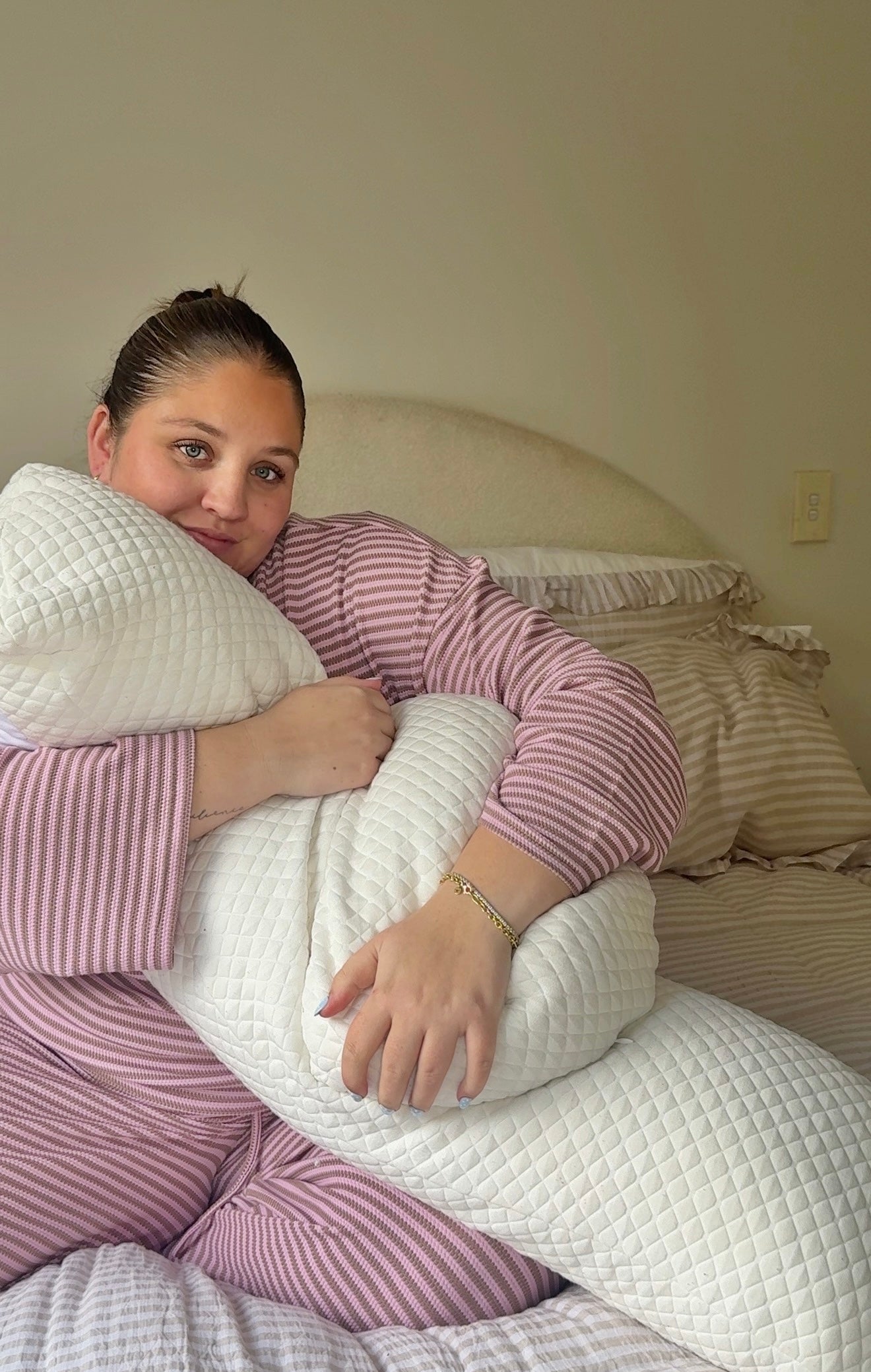 A woman in pink and white striped pajamas sits on a bed holding a white quilted pregnancy pillow.