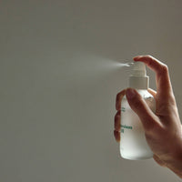 Hand holding a white spray bottle with a mist being released against a plain background